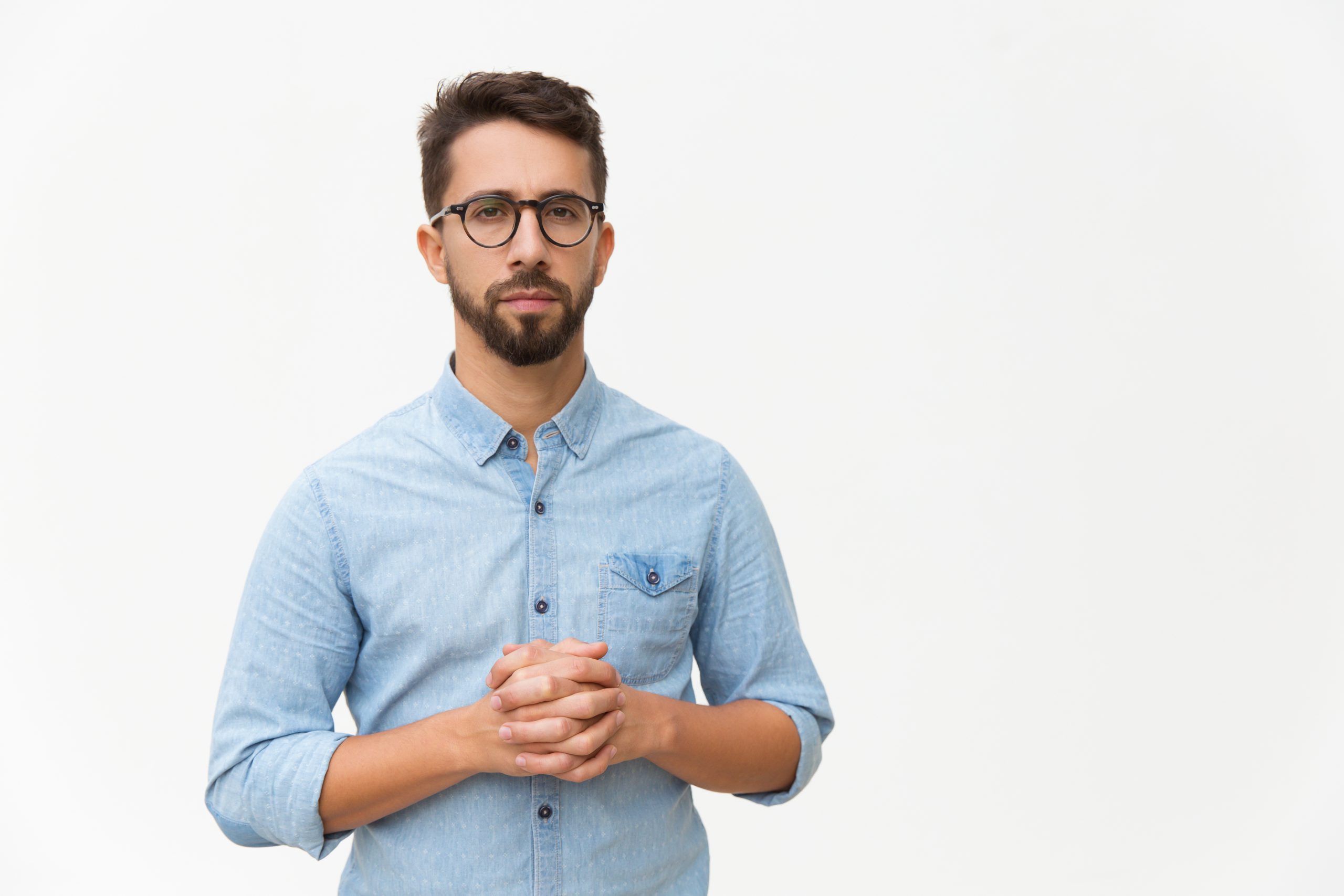 Front of serious confident man with clasped hands looking at camera. Handsome young man in casual shirt and glasses standing isolated over white background. Male portrait concept